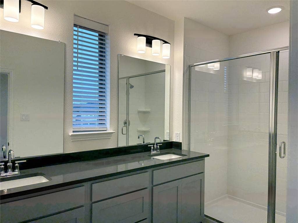 2528 Brunswick Way, Allen, TX 75013 Dual-sink vanity with dark stone-finish countertop, crisp shaker-style cabinetry, glass-enclosed shower, and sleek modern wall-mounted lighting