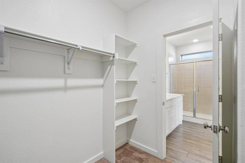 135 Lost Maples Way, Waxahachie, TX 75165 Walk-in closet featuring white built-in shelving and a garment rod, with a doorway revealing a bathroom equipped with wood-finish flooring, a white cabinet vanity, and a tiled shower stall