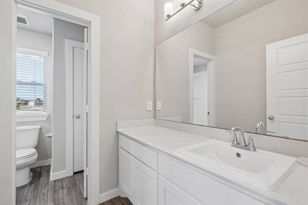 135 Lost Maples Way, Waxahachie, TX 75165 Bathroom featuring a white shaker-style vanity with a composite countertop, an undermount sink, a full-width wall mirror, and wood-finish flooring