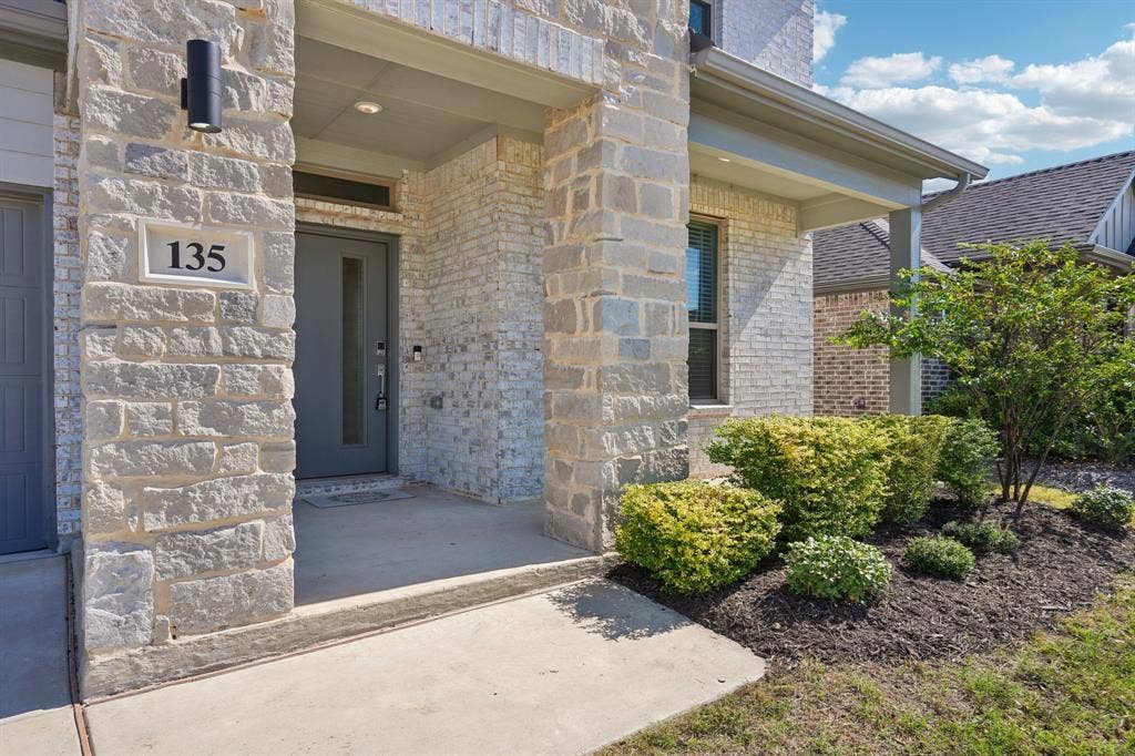 135 Lost Maples Way, Waxahachie, TX 75165 Covered front porch featuring stacked stone accents, gray-toned masonry, a modern entry door with a vertical glass insert, and wall-mounted exterior cylinder lighting