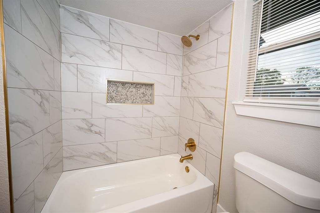 2406 Cliff Teen Court, Dallas, TX 75233 Bathtub enclosure featuring large-format stone-patterned wall tile, a recessed mosaic-tiled soap niche, and brass-finish plumbing fixtures