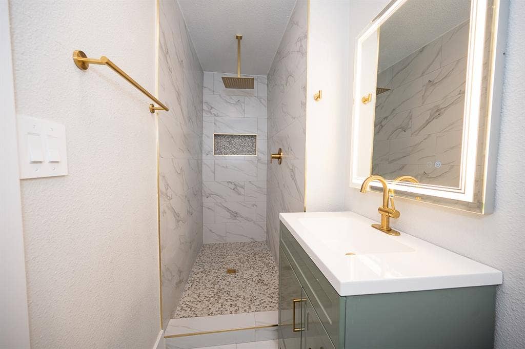 2406 Cliff Teen Court, Dallas, TX 75233 Walk-in shower featuring tiled walls with stone-pattern detail, pebble-tile flooring, and a rainfall showerhead; vanity with a white countertop and gold-finish hardware; backlit mirror