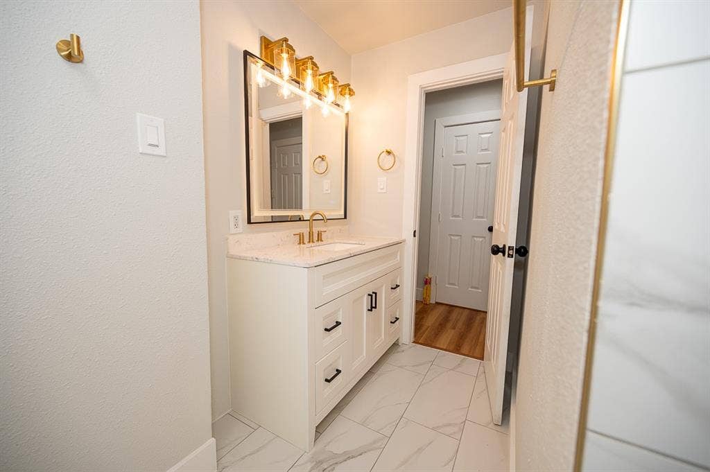 2406 Cliff Teen Court, Dallas, TX 75233 White vanity cabinet with stone-composite countertop, rectangular undermount sink, brass-finish faucet, four-light vanity fixture, and large-format tile flooring