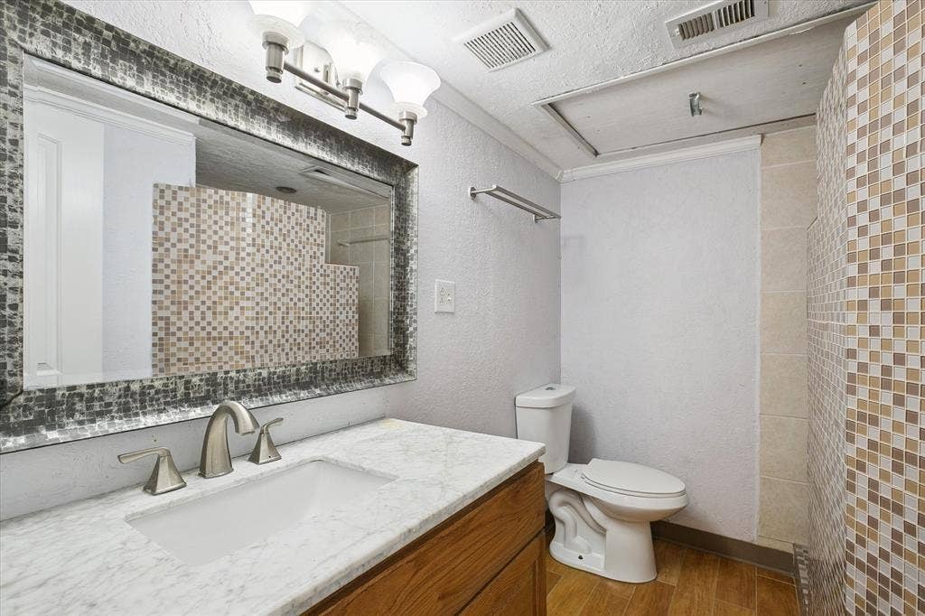 513 Towne House Lane, Richardson, TX 75081 Bathroom featuring a vanity with a light-toned stone countertop, undermount sink, multi-tone mosaic tile backsplash, framed wall mirror, and wood-finish flooring