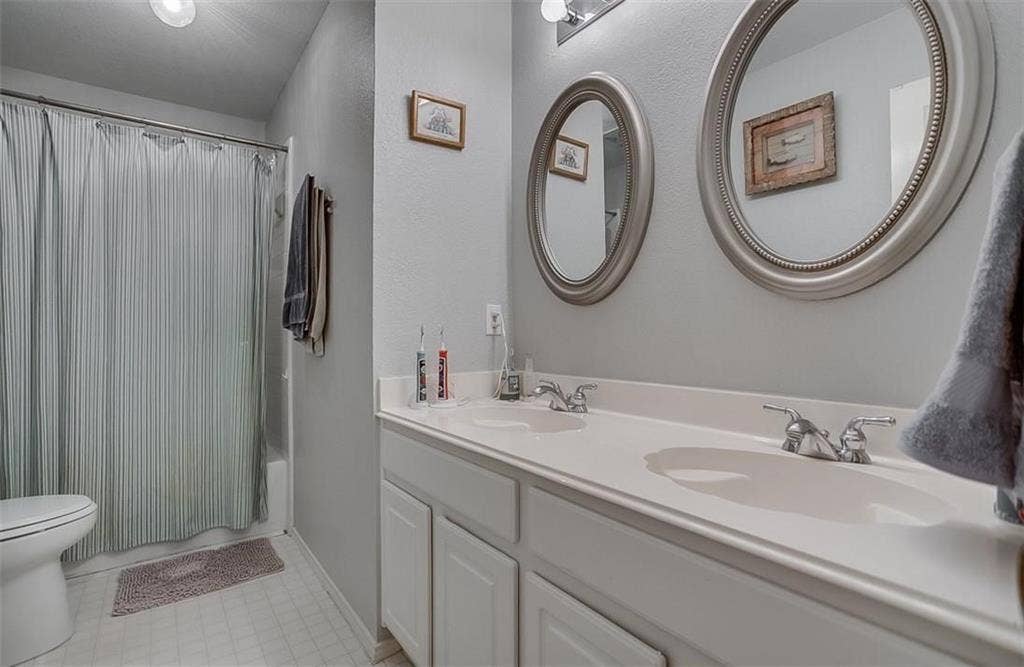 1307 Capstan Drive, Allen, TX 75013 Dual-sink vanity with a solid-surface countertop, two oval wall-mounted mirrors, chrome plumbing fixtures, and a shower-tub combination with an enclosure