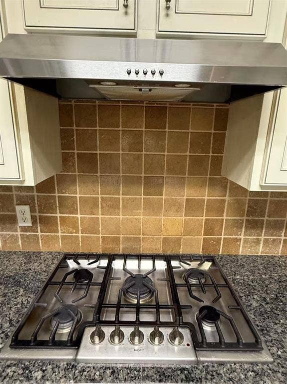 1307 Capstan Drive, Allen, TX 75013 Five-burner stainless steel gas cooktop with heavy-duty grates, matching stainless steel range hood, natural stone tile backsplash, and dark stone-surfaced countertop with light-toned cabinetry