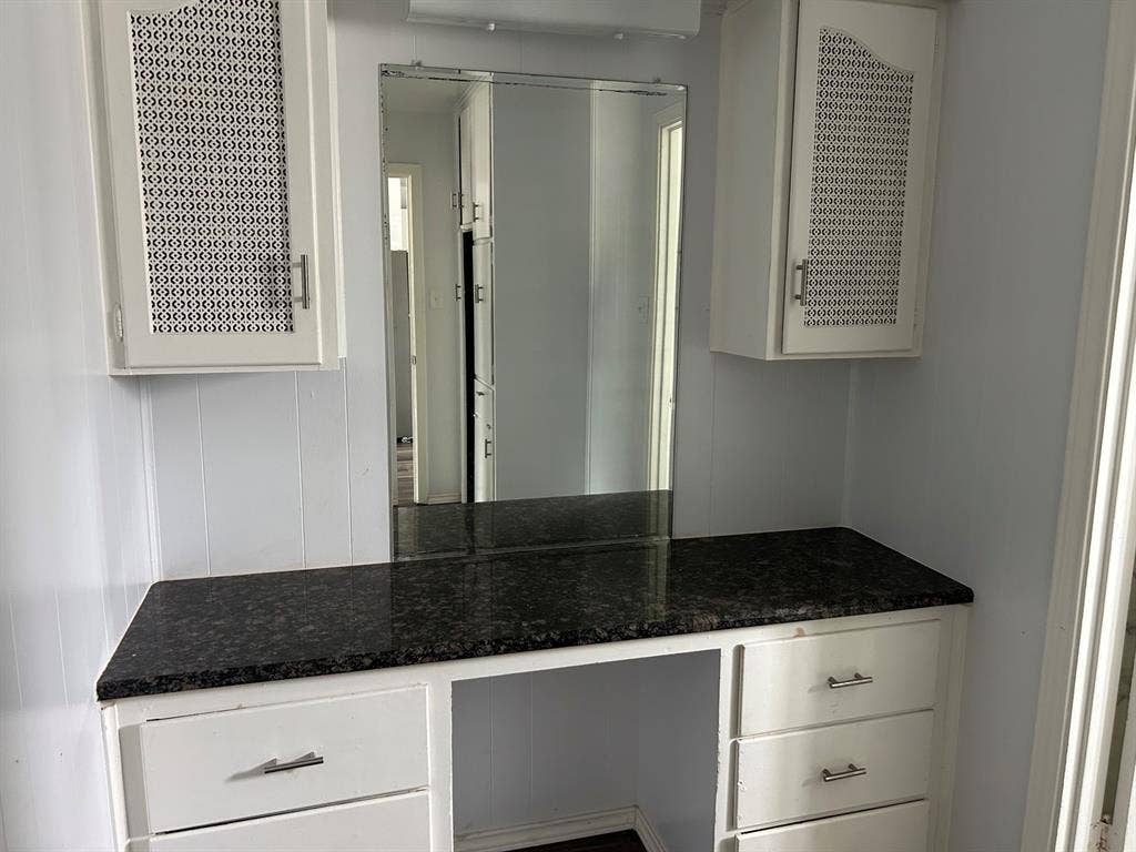 Built-in vanity featuring white cabinetry, dark stone-finish countertop, upper wall-mounted storage cabinets with decorative mesh panels, and a full-height mirror
