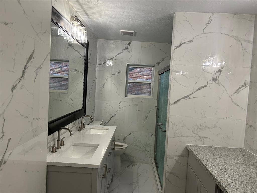 Bathroom featuring floor-to-ceiling large-format veined tile, double-sink vanity with a solid surface countertop, framed wall mirror, and glass-enclosed shower
