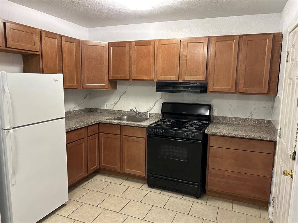 Kitchen featuring wood cabinetry, stone-finish countertops, full-height backsplash, neutral tile flooring, and integrated black appliance package