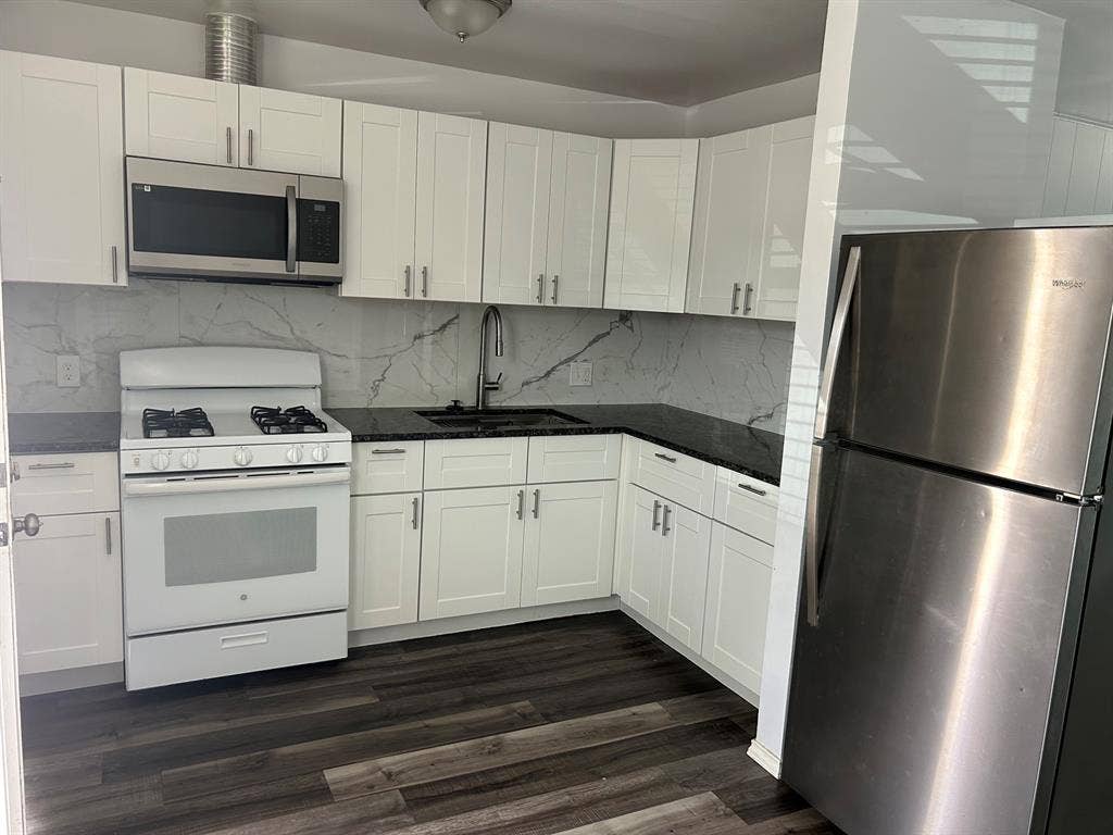Kitchen featuring white shaker-style cabinetry, dark stone countertops, a full-height stone-pattern backsplash, stainless steel appliances, and wood-finish flooring