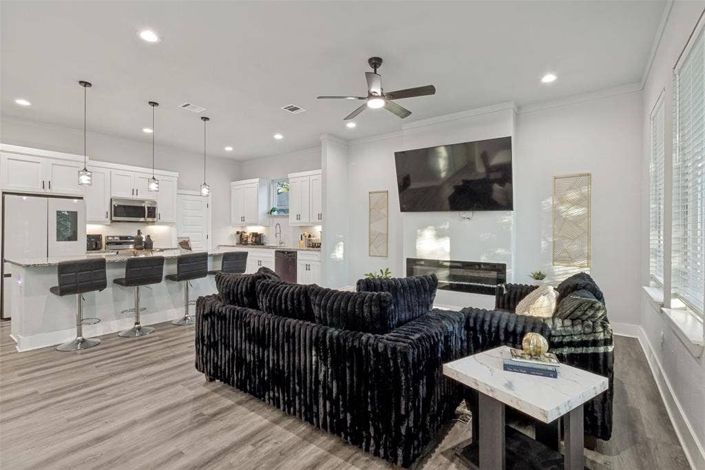 7909 Ivory Lane, Dallas, TX 75216 Open concept kitchen featuring white shaker-style cabinetry, an expansive kitchen island with a neutral countertop, built-in stainless steel appliances, recessed lighting, and wood-finish flooring throughout, complemented by a floor-to-ceiling fireplace surround