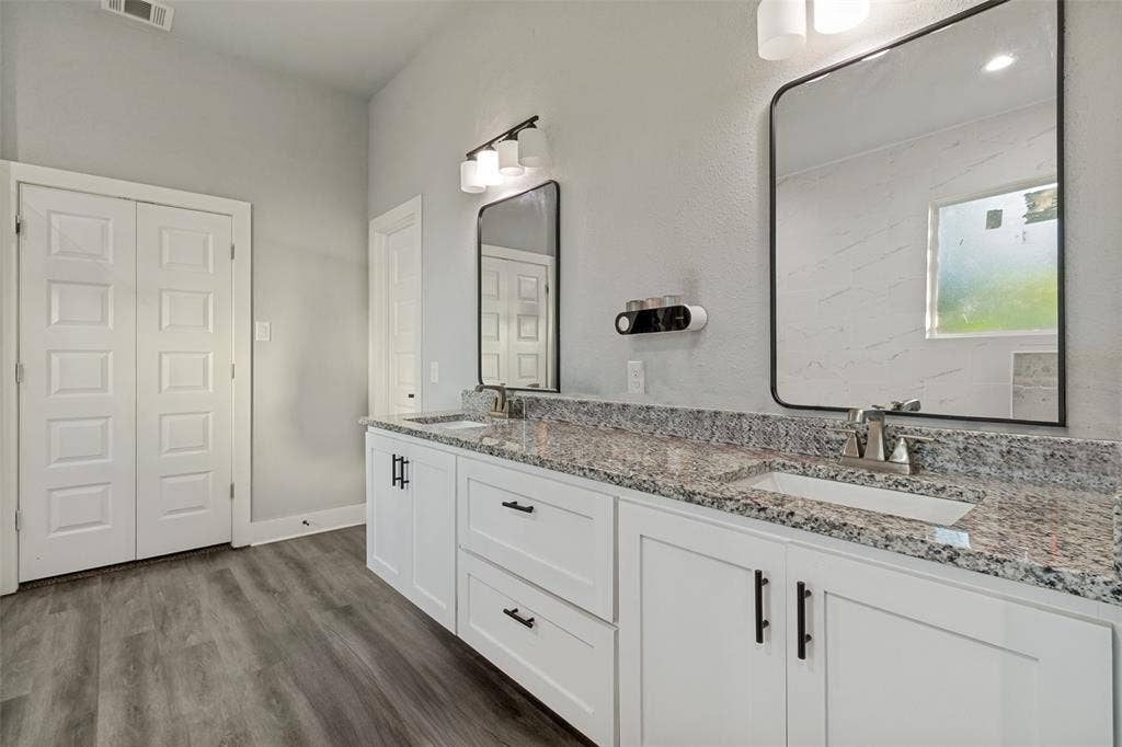 7909 Ivory Lane, Dallas, TX 75216 Dual-vanity bathroom featuring white cabinetry with matte black hardware, speckled stone-patterned countertops, dual rectangular under-mount sinks, wood-finish flooring, and framed mirrors