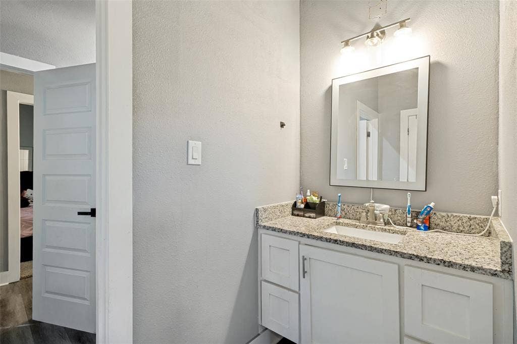 7909 Ivory Lane, Dallas, TX 75216 White shaker-style vanity featuring a stone-finish countertop, integrated rectangular sink, framed wall mirror, and three-bulb vanity light fixture