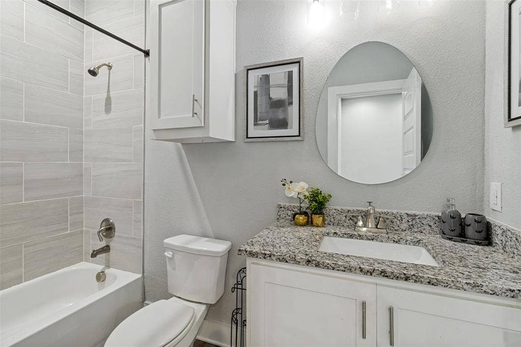 7909 Ivory Lane, Dallas, TX 75216 Bathroom featuring a stone-composite countertop, undermount sink, white cabinetry with brushed-nickel hardware, an oval wall-mounted mirror, and tile shower surround
