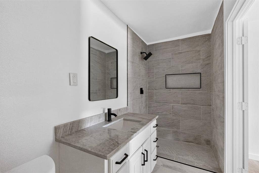 1831 Highland Street, Mesquite, TX 75149 Bathroom featuring a white vanity with stone-finish countertop, undermount sink, matte black hardware, rectangular mirror, and walk-in shower with large-format tile surround and built-in recessed niche