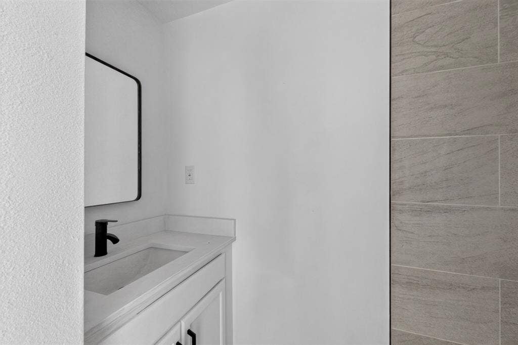 1831 Highland Street, Mesquite, TX 75149 Built-in vanity featuring a solid-surface countertop, rectangular integrated sink, matte black faucet, and rectangular wall-mounted mirror, accented by a textured stone-finish tile wall