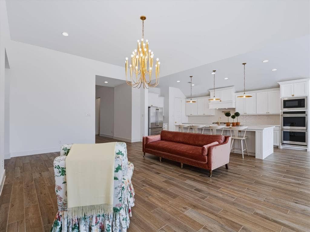 1103 Sandgate Drive, Forney, TX 75126 Open-concept interior featuring wood-finish tile flooring, white shaker-style cabinetry, a built-in double oven, quartz-topped kitchen island, and a multi-tiered gold-tone chandelier