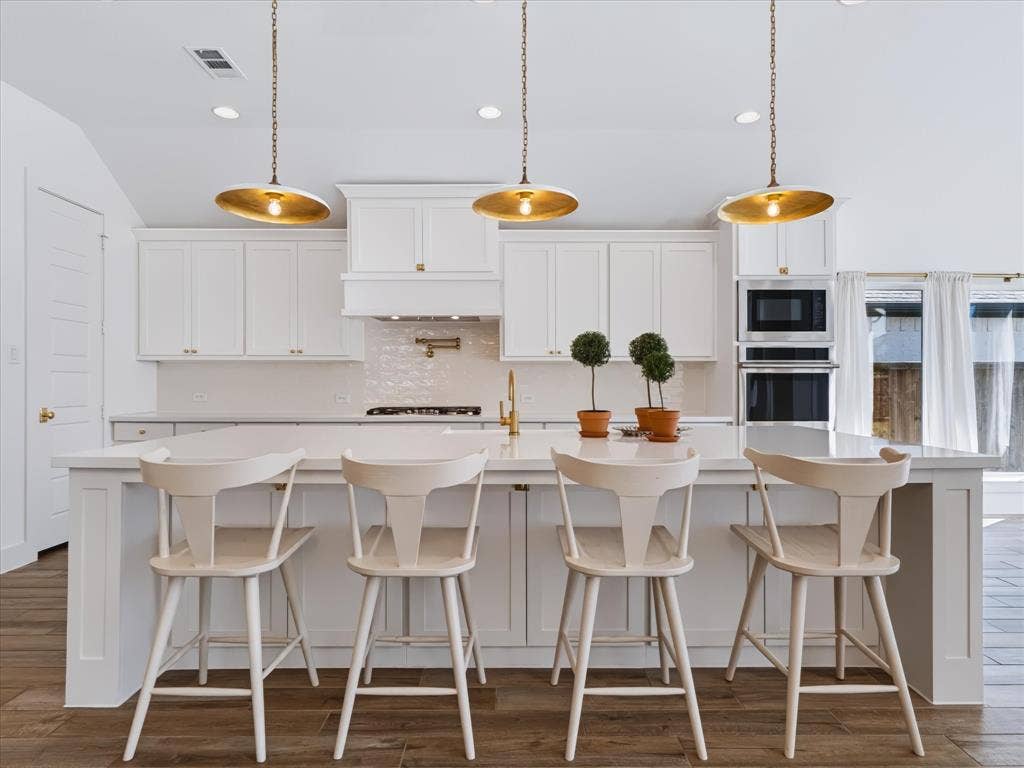1103 Sandgate Drive, Forney, TX 75126 Kitchen featuring an oversized central island with solid-surface countertop, white shaker-style cabinetry, stainless steel built-in double ovens, gold-toned hardware, and three pendant light fixtures