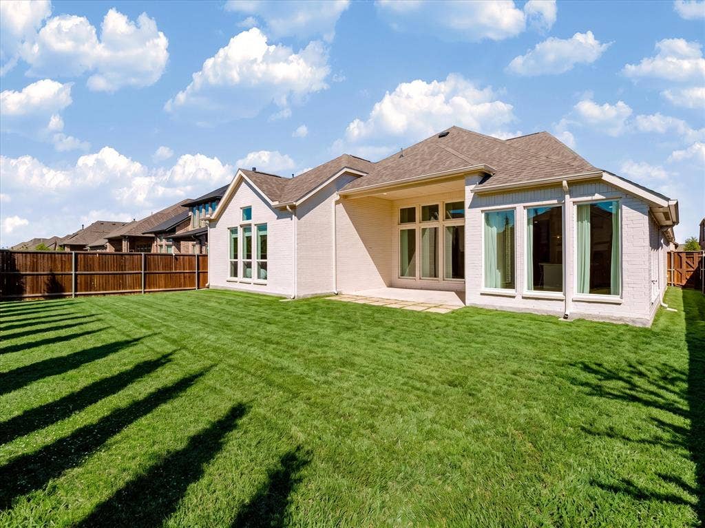 1103 Sandgate Drive, Forney, TX 75126 Light-colored brick exterior featuring a covered patio, expansive manicured lawn, full-height multi-pane windows, and wood-style perimeter fencing