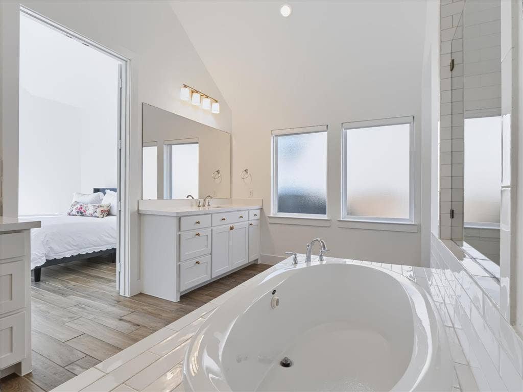 1103 Sandgate Drive, Forney, TX 75126 Vaulted ceiling bathroom featuring a white dual-sink vanity, recessed oval soaking tub, subway tile surround, and wood-finish flooring