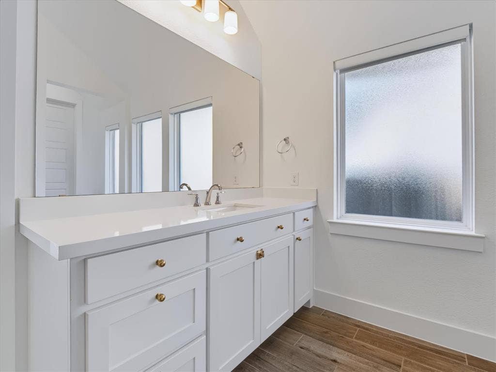 1103 Sandgate Drive, Forney, TX 75126 White vanity with a solid-surface countertop, under-mount sink, brushed gold-finish hardware, full-width mirror, and wood-finish flooring
