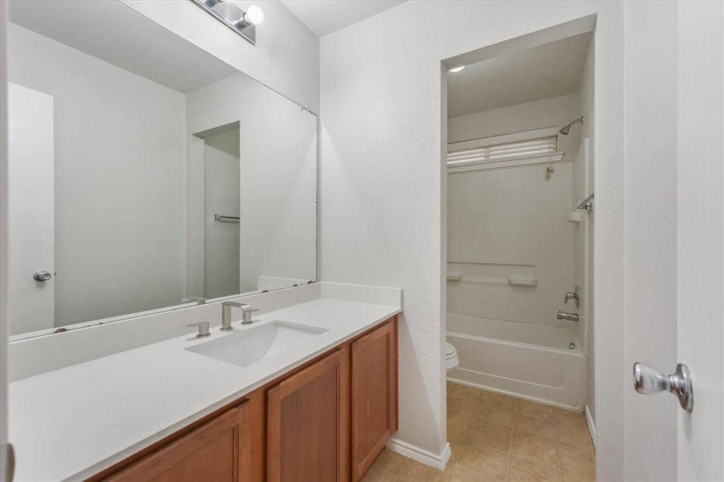 2004 Athabasca Falls Drive, Anna, TX 75409 Full-width vanity with a solid-surface countertop, integrated rectangular sink, wood-finish cabinetry, and oversized wall-to-wall mirror, featuring an enclosed tub and shower combination with tile surround