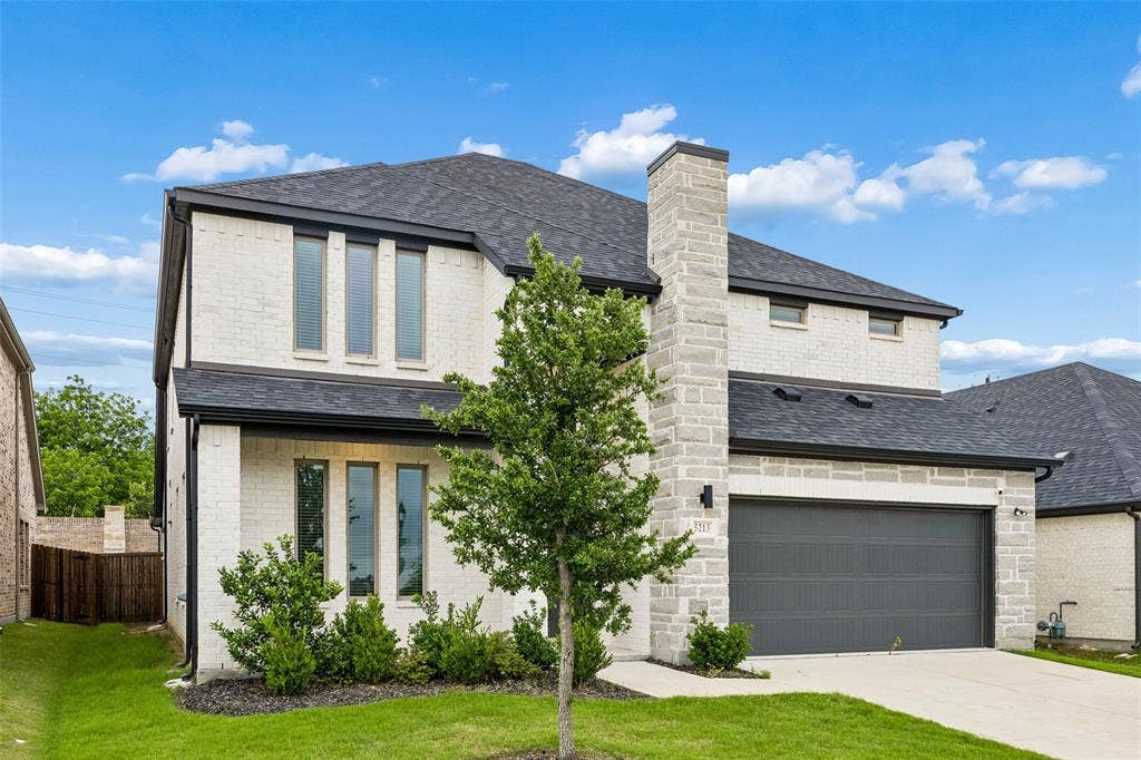 Two-story exterior featuring a white brick and stone facade, a multi-gabled asphalt shingle roof, a two-car garage with a paneled door, and a covered entryway