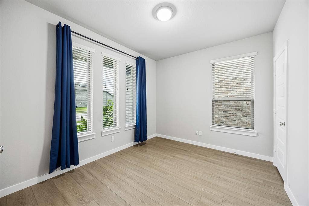 Neutral interior space featuring wood-finish flooring, four windows with integrated blinds, and a flush-mount overhead light fixture