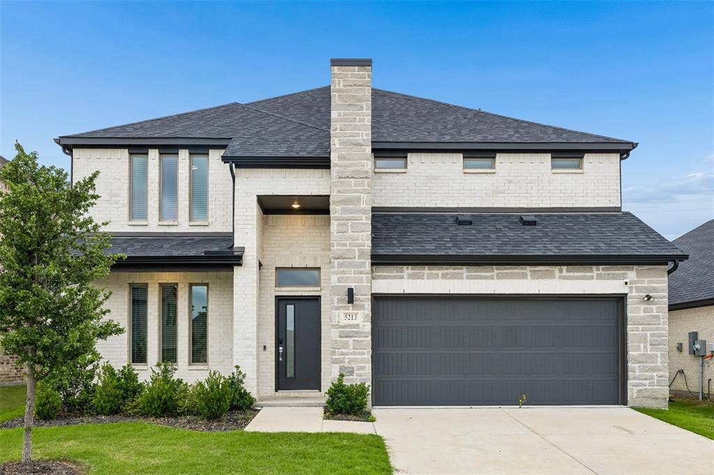 Two-story exterior featuring white brick and stone facade, dark-toned multi-gabled roof, integrated two-car garage, and vertical window panels
