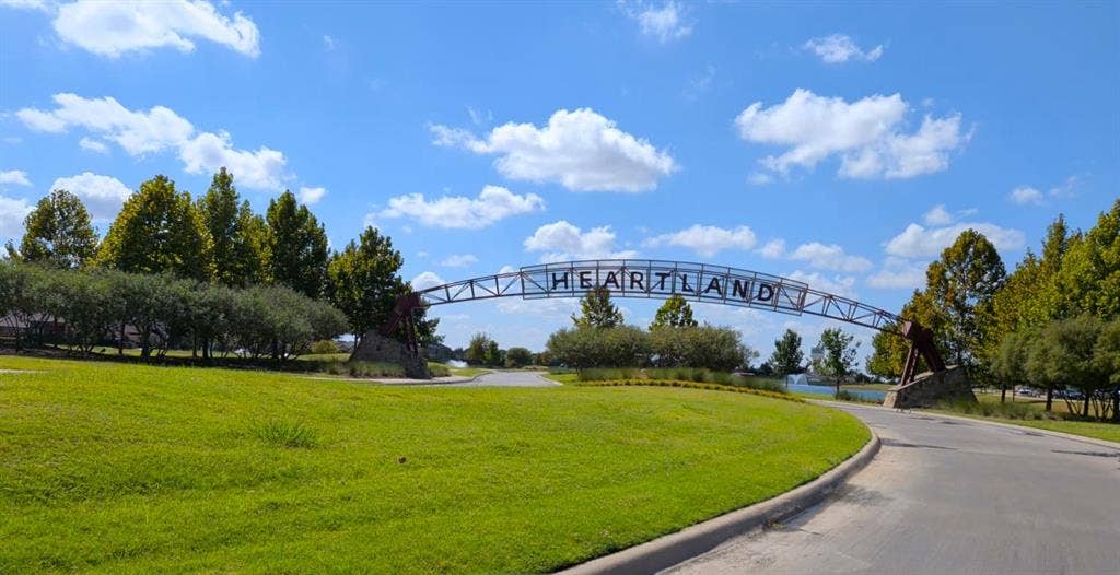 Community entrance featuring an arched metal signage structure, stone-accented support pillars, expansive manicured turf, and mature landscaping