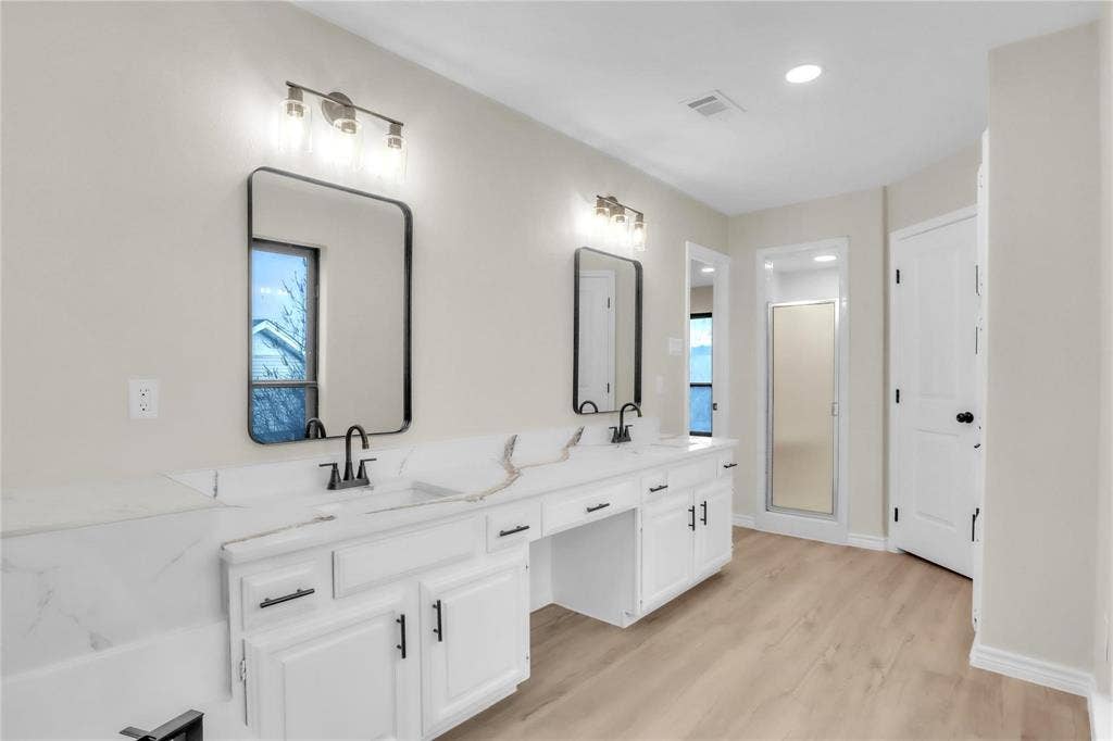 600 Flintridge Court, Irving, TX 75038 Dual-sink vanity with white cabinetry, light-toned countertop, rectangular wall-mounted mirrors, and matte black fixtures, paired with wood-finish flooring and a walk-in shower enclosure