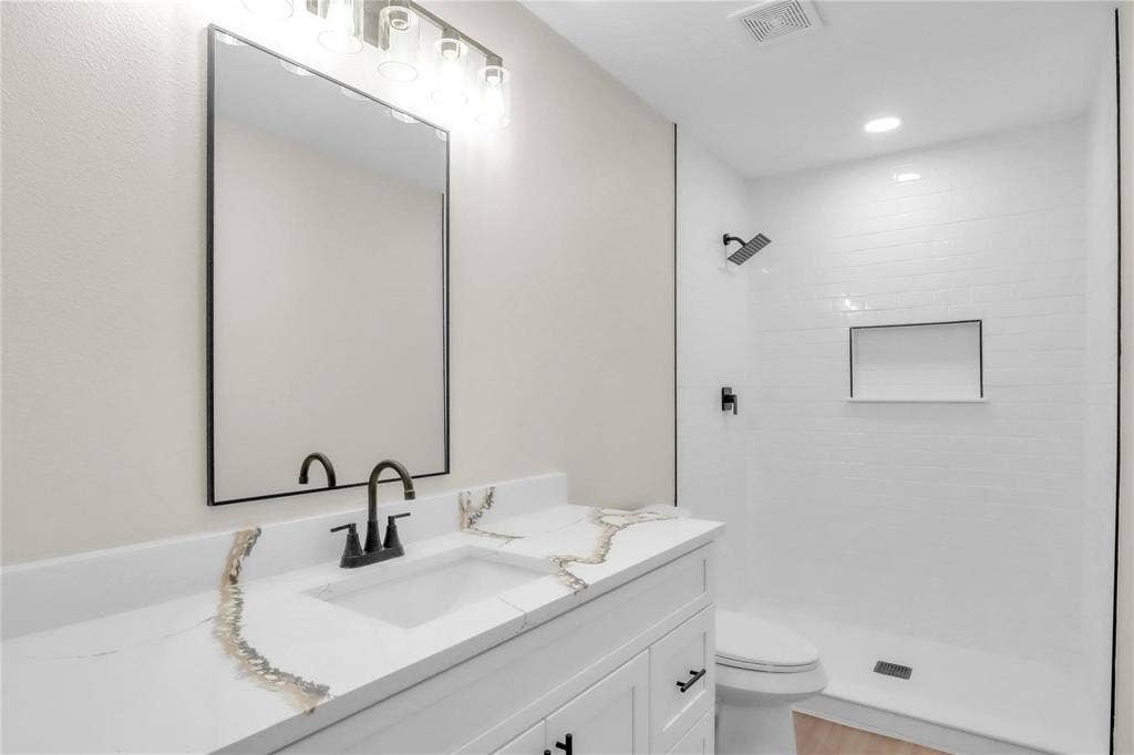 600 Flintridge Court, Irving, TX 75038 Bathroom featuring a white vanity with a stone countertop, rectangular undermount sink, black metal hardware, framed mirror, and a walk-in shower with white wall tile and a built-in recessed niche