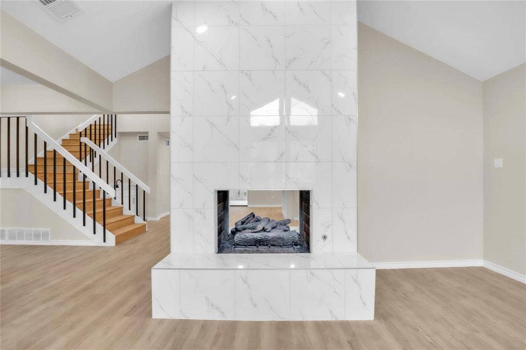 600 Flintridge Court, Irving, TX 75038 Double-sided fireplace with large-format white stone-finish tile surround and hearth, featuring a staircase with white balusters, black handrail, and wood-finish treads, set against high vaulted ceilings and light wood-finish flooring