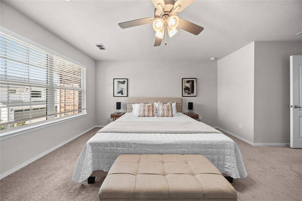 Neutral-toned room featuring wall-to-wall carpeting, large windows with horizontal blinds, and a central ceiling fan with multi-light fixture