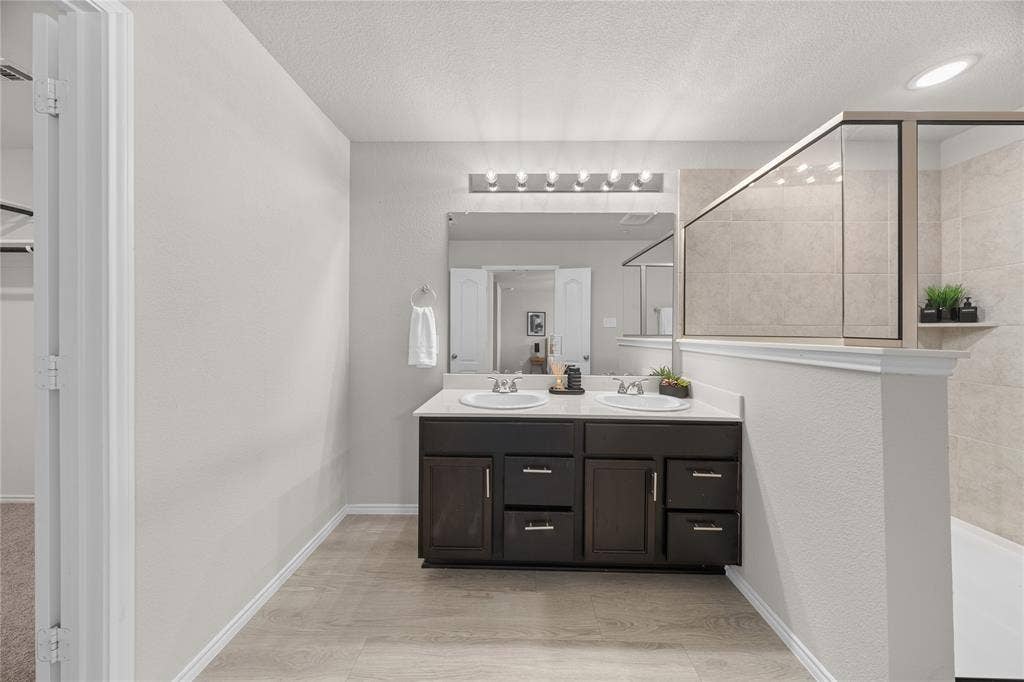 Bathroom featuring a dual-sink vanity with dark cabinetry, light-toned countertop, large wall-mounted mirror, multi-bulb vanity lighting, and a tiled shower enclosure