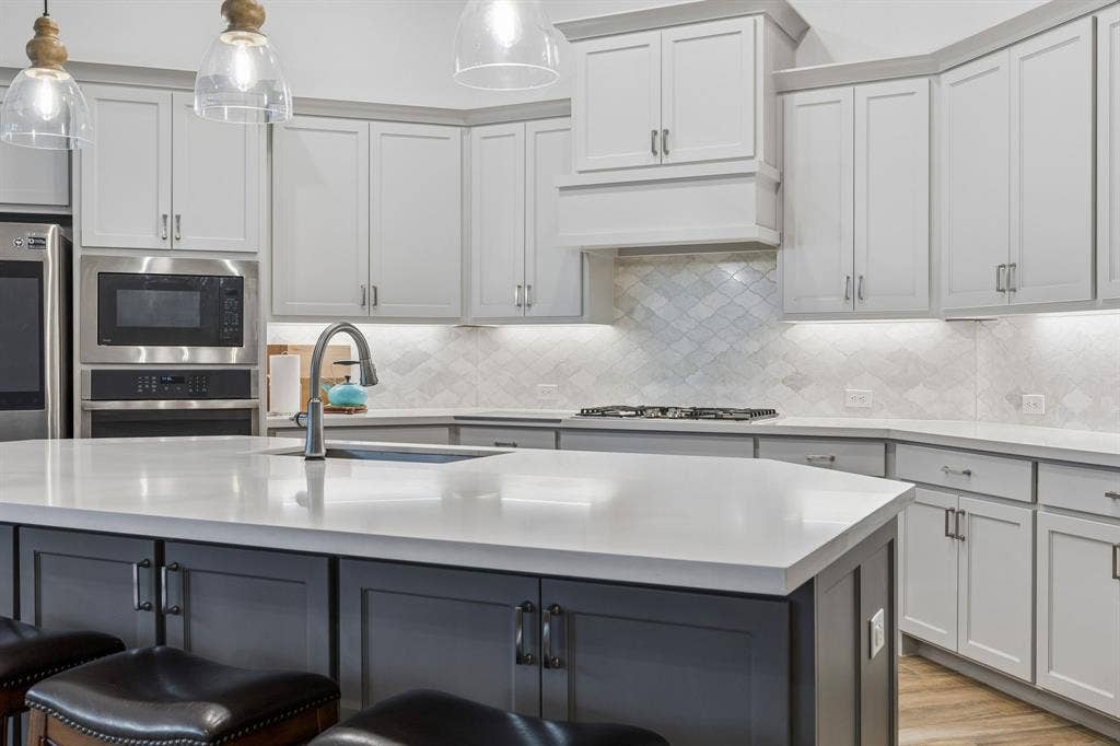 Kitchen featuring light gray shaker-style cabinetry, quartz countertops, a multi-tonal diamond pattern tile backsplash, stainless steel built-in microwave and oven, and a center island with contrasting charcoal-toned cabinetry