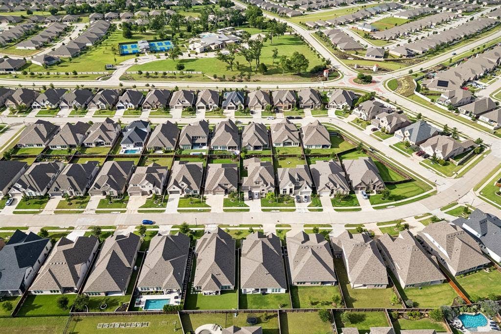 Aerial perspective of a planned community featuring uniform residential roofing, paved street infrastructure, landscaped green space, and a community recreation area with tennis courts