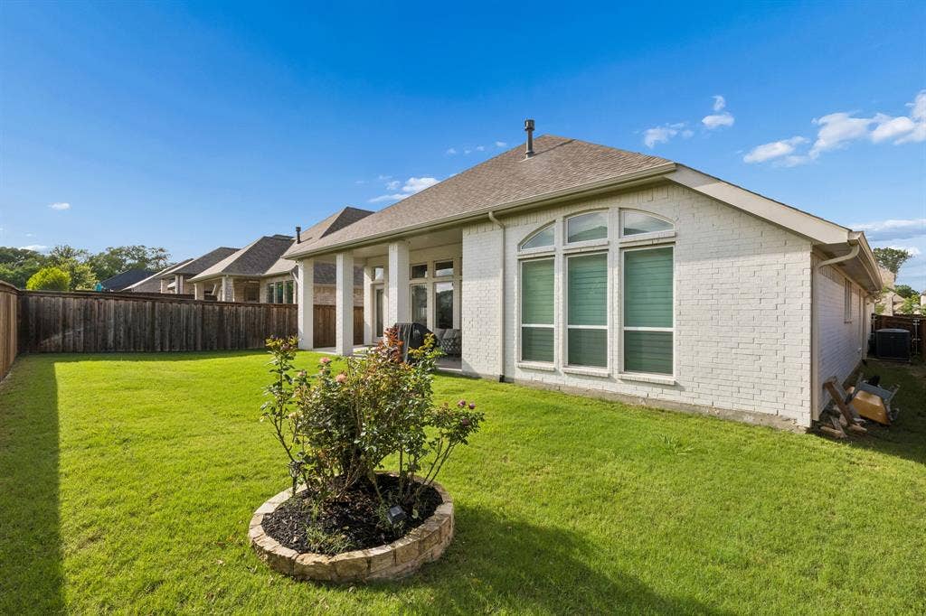Rear exterior featuring a white brick facade, covered patio, manicured lawn, stone-bordered garden bed, and wood privacy fencing