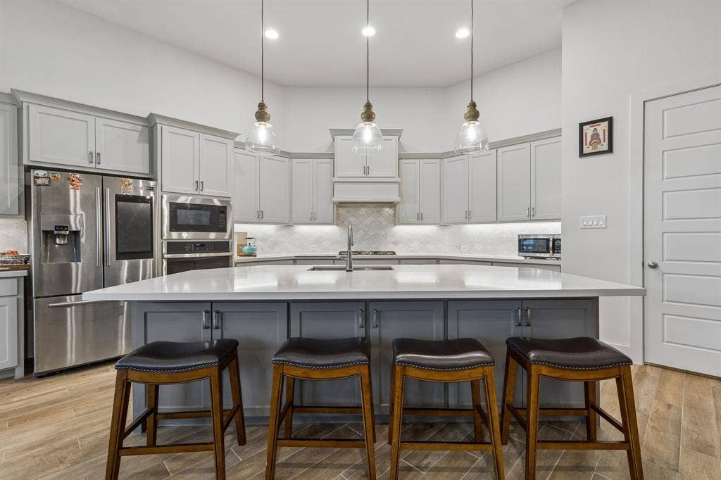 Kitchen featuring a central island with a solid surface countertop, light gray shaker-style cabinetry, stainless steel built-in appliances, herringbone pattern tile backsplash, and three glass pendant lights