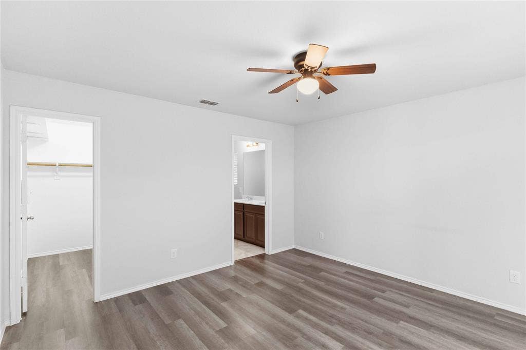 Room featuring wood-finish flooring, a central ceiling fan with integrated lighting, and access to a walk-in closet and a separate bathroom with dark wood-toned cabinetry