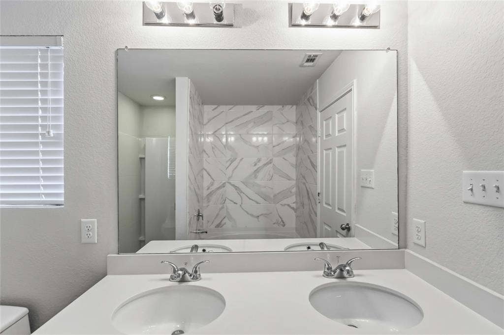 Dual-sink vanity with white surface, expansive wall-mounted mirror, polished chrome fixtures, and tiled tub surround