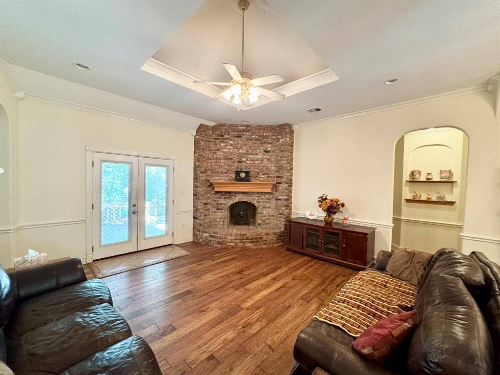3700 NE County Road 0095, Corsicana, TX 75109 Vaulted ceiling with crown molding, corner brick fireplace featuring a wooden mantel, wood-finish flooring, double glass panel exterior doors, and a built-in arched wall niche with shelving