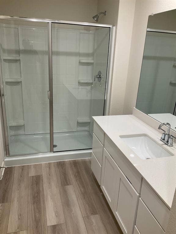 325 Stone Court, Princeton, TX 75407 Bathroom featuring wood-finish flooring, white vanity cabinetry, a rectangular undermount sink with a quartz countertop, a large wall-mounted mirror, and a walk-in shower with a sliding glass door and integrated shelving