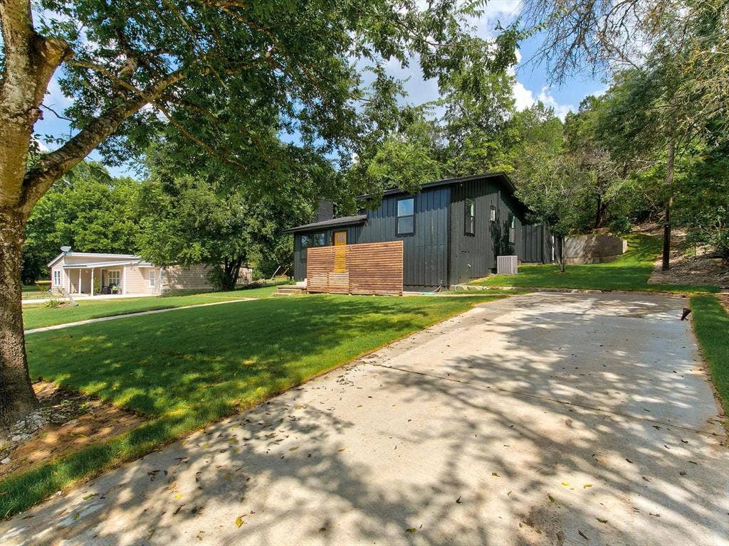 Exterior featuring black vertical siding, natural wood privacy screen, concrete driveway, and expansive lawn with mature trees