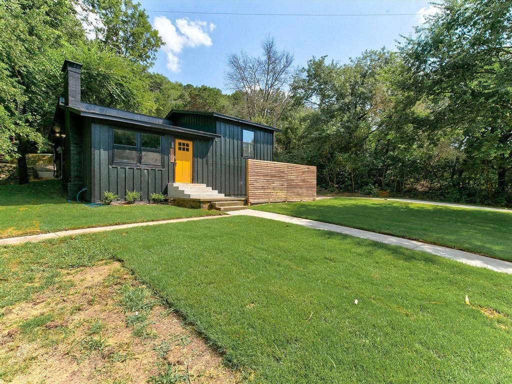 Exterior featuring vertical dark-tone siding, a brick chimney, a contrasting yellow front door, and wood-slat privacy screening