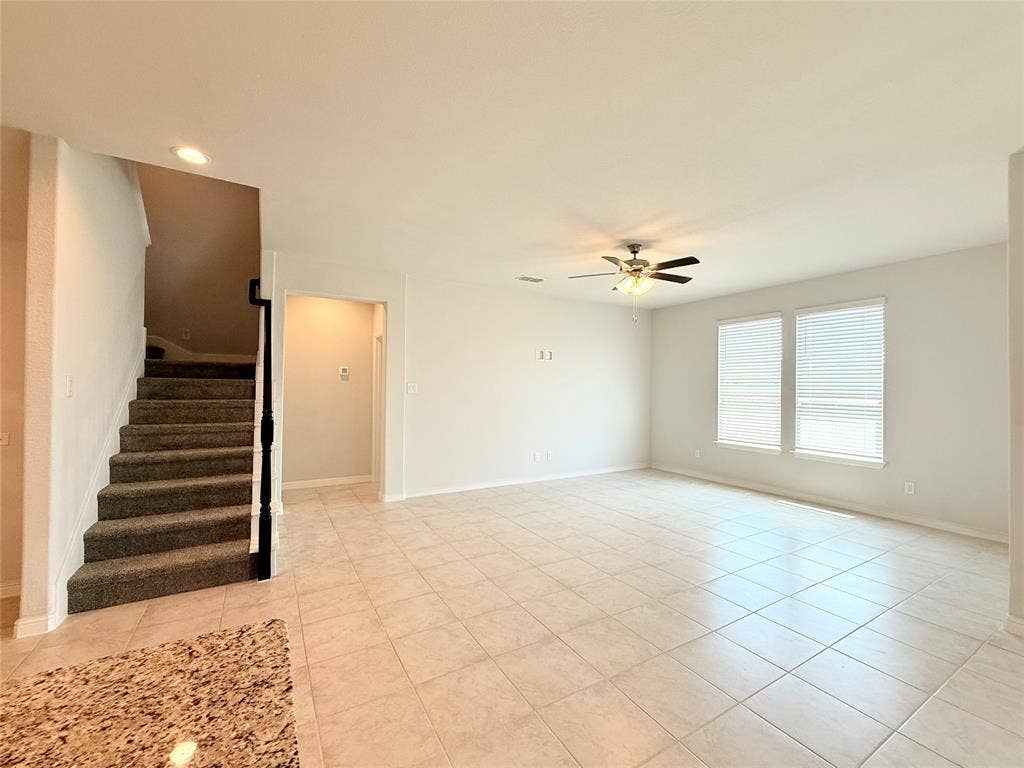 Open interior space featuring light-toned tile flooring, recessed ceiling lighting, a multi-blade ceiling fan, and a staircase with carpeted treads