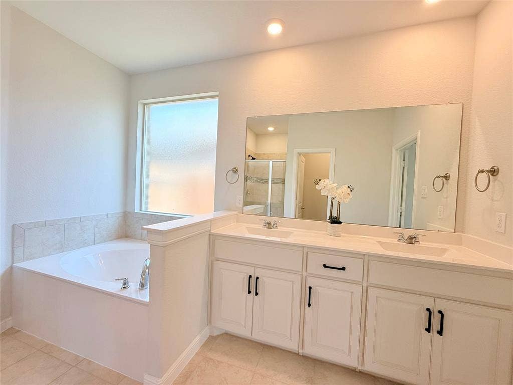 Double vanity with white cabinetry, rectangular under-mount sinks, and large wall-to-wall mirror, featuring a built-in soaking tub with a tile surround and recessed lighting