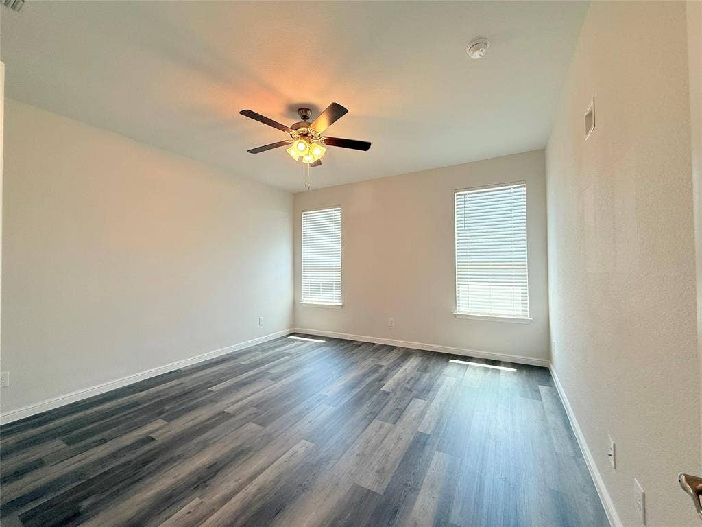 Neutral-toned room featuring wood-finish flooring, two windows with horizontal blinds, and a ceiling fan with integrated lighting