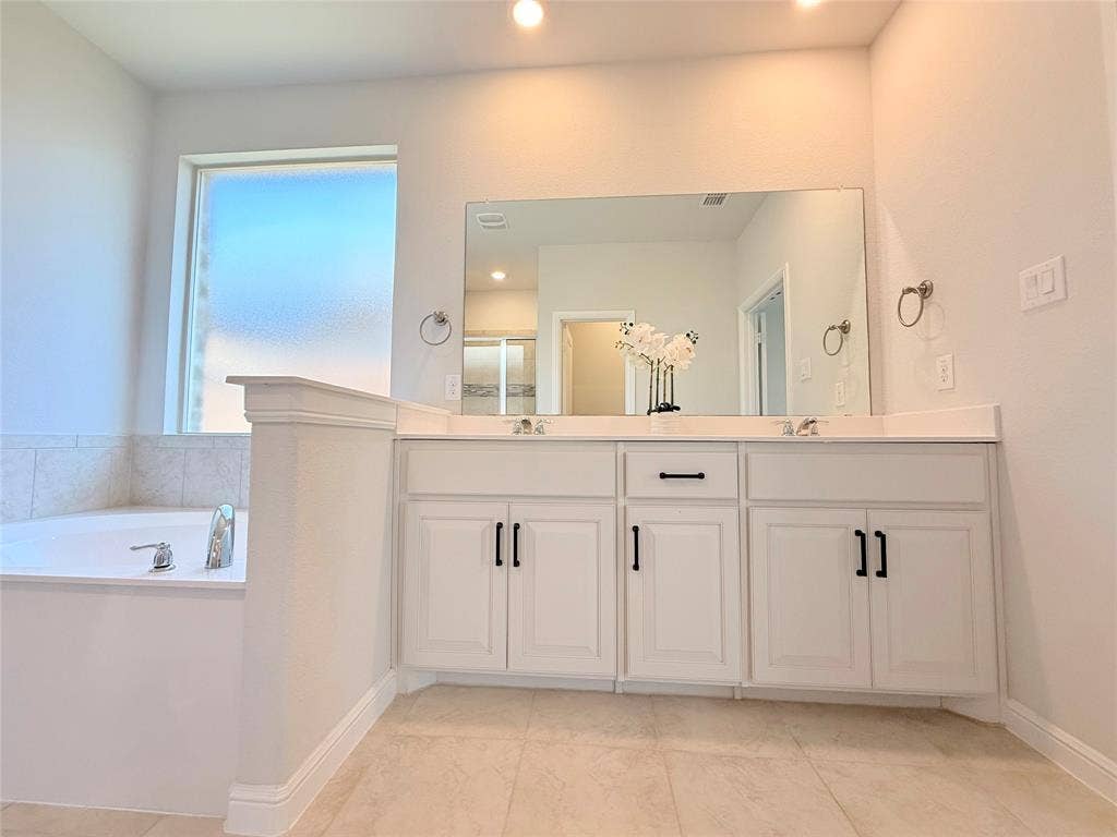 Bathroom featuring a soaking tub with tile surround, white cabinetry, dual-sink vanity, large rectangular mirror, and matte black hardware