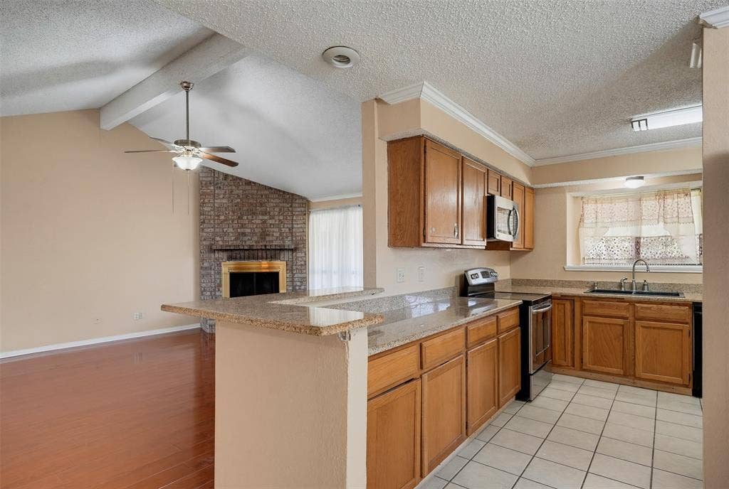 Kitchen featuring wood cabinetry, stone-finish countertops, tile flooring, and stainless steel appliances, with a vaulted ceiling and brick fireplace beyond the breakfast bar